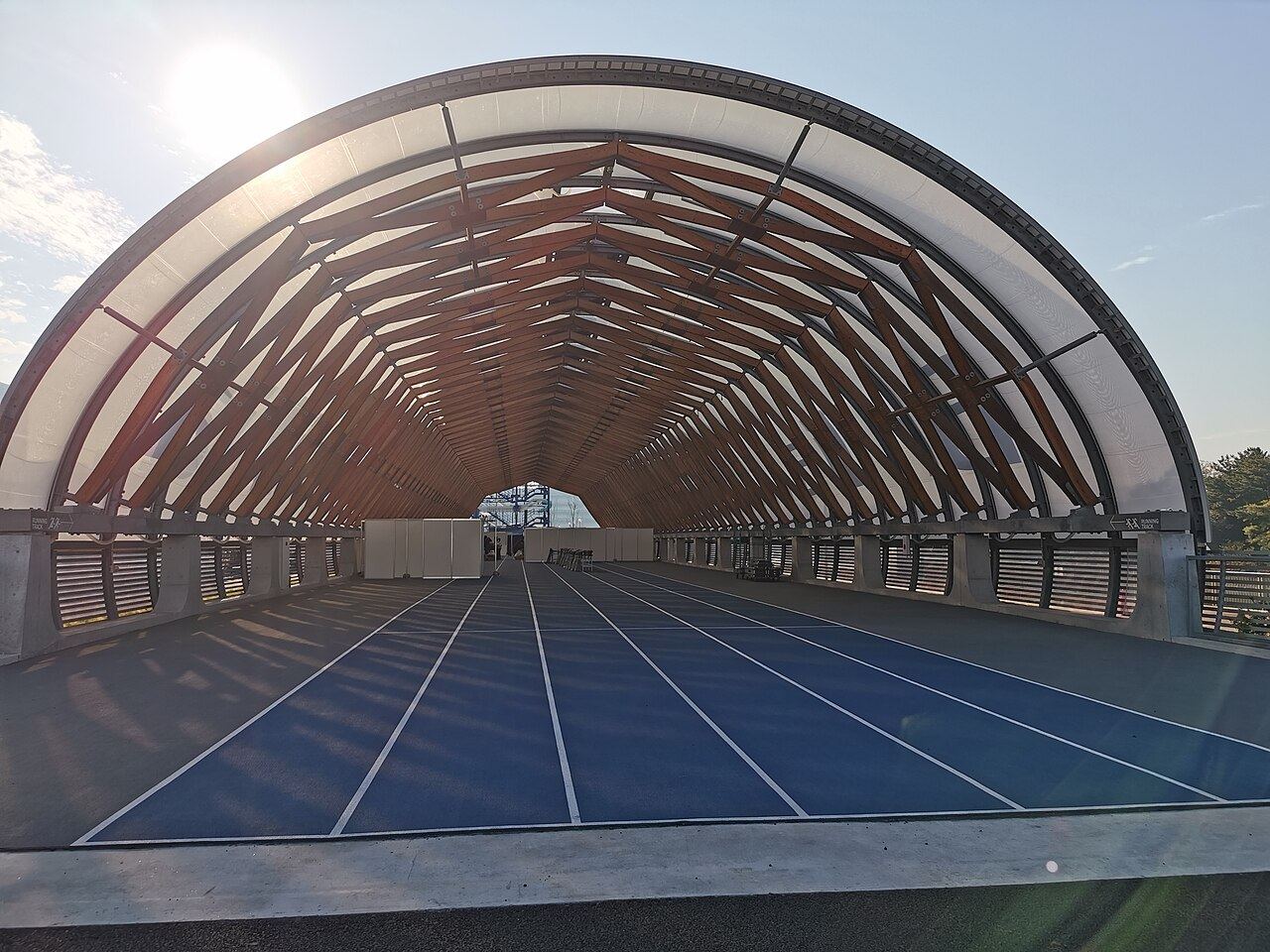 Piste d'athlétisme bleue et vide sous une charpente en arc de bois, éclairée par le soleil du matin