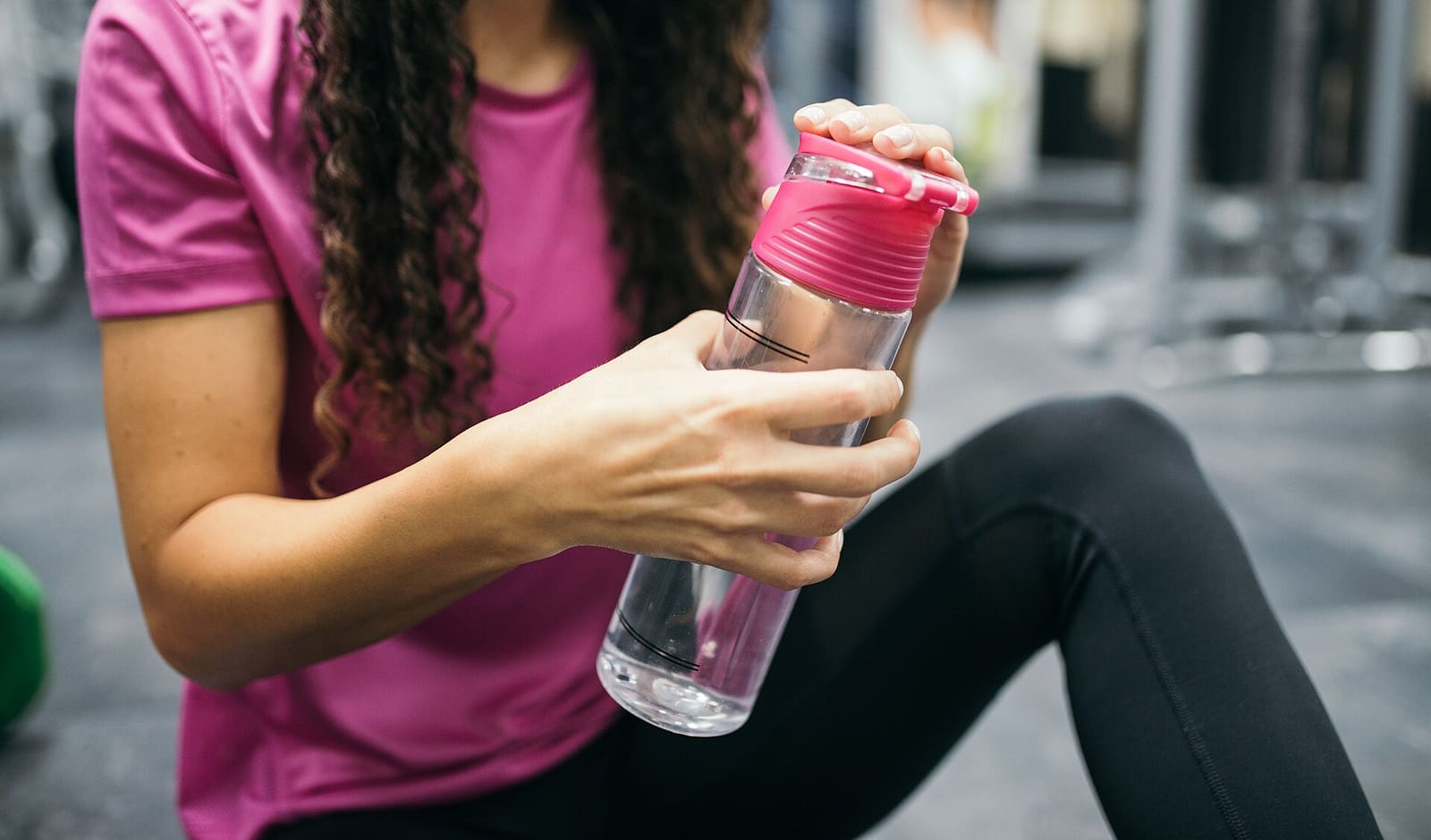 Une femme assise au sol d'une salle de sport ouvre une gourde d'eau transparente