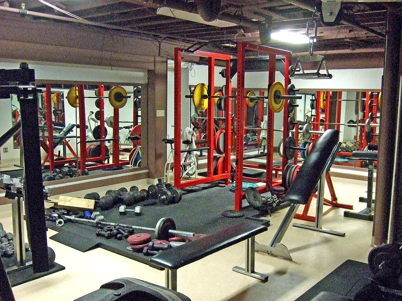 Salle de musculation vide, avec des cages à squat rouges, des barres chargées de disques et des haltères au sol