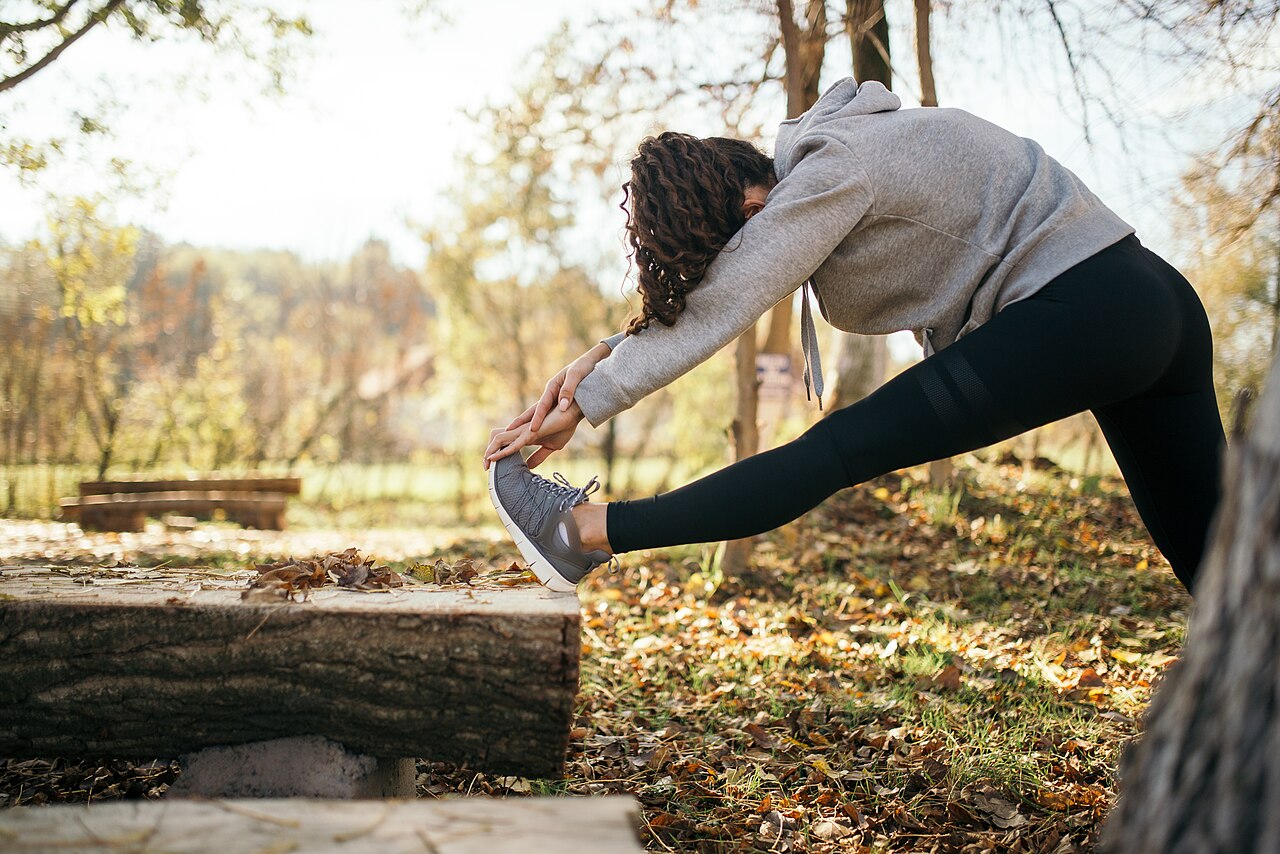 Femme en sweat gris qui étire l'arrière de sa cuisse, le pied posé sur un tronc couché, dans un sous-bois d'automne